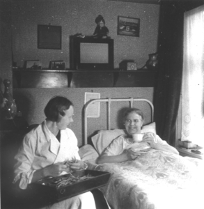 876919 Afbeelding van een Zuster Augustines (in opleiding?) die als gezinshulp werkt, zittend naast het bed van een ...